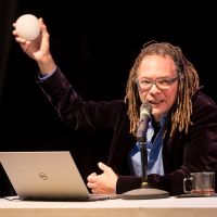 Thomas F DeFrantz sits at a desk with his laptop and is holding up a ball.